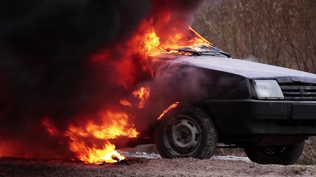 Huge red flame of fire with massive matt black smoke completely burns rusty dirty old dark dusty violet car which is left on deserted grey road at some suburb area on cold cloudy winter day.