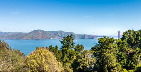 A panoramic of the Golden Gate Bridge and Highlands from Lincoln Park across the entrance to the bay. Trees are in the foreground. Blue sky.