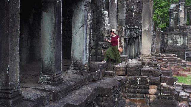 Young woman in hat and sunglasses is walking on ruins of of Bayon Temple, a Khmer temple at Angkor in Cambodia built in the late 12th or early 13th century and richly decorated