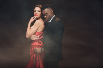 african american man embracing beautiful woman in red dress on dark with smoke