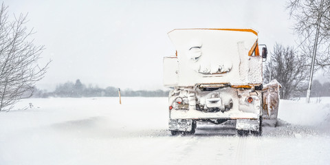 Orange plough truck on snow covered road, gray sky and trees in background, view from back - winter...