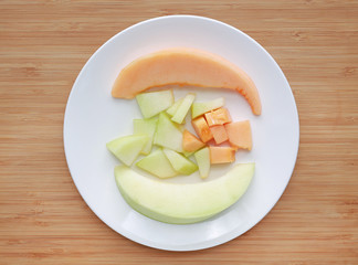 Fresh sliced of green and orange cantaloupe melon on white plate against wooden board background. 