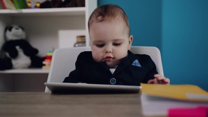 Close up view of extremely adorable baby boy in business suit holding tablet in his hands while sitting by the table in modern cozy office. Baby boss concept, modern gadgets, childhood. Child�s