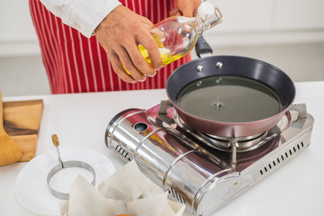 A man wearing a white shirt and red apron pouring oil into pan in a white kitchen.	