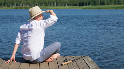 a man in a hat,sitting on the pier,looking into the distance, lying next to a fishing rod, against the lake