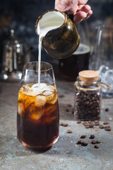 Ice coffee cold summer drink in a tall glass with pouring cream and coffee beans on a stone background.