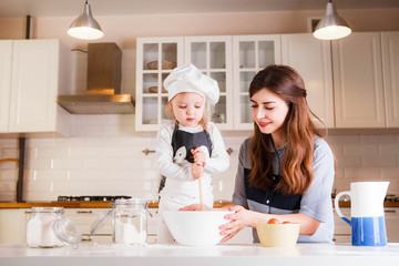 The little daughter in the chef's hat and apron and her mother prepare baking in the bright, classic kitchen.