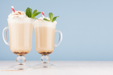Cappuccino coffee in two elegant glass with whipped cream, cocoa powder, green mint and red straw on white table and soft light blue wall, copy space.