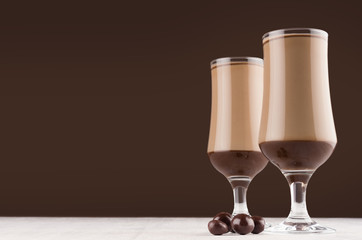 Season coffee beverage in two glass with chocolate balls candies on dark brown wall and white wooden table, copy space.