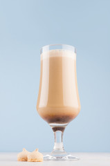 Summer coffee cocktail with cream and biscuits for breakfast in transparent glass in soft pastel blue interior on white wood table, vertical.