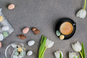 A cup of aromatic coffee with froth over a gray background, white tulips and chocolates for Easter. Copy space, flat lay.