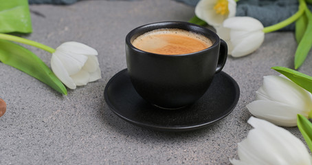 A cup of aromatic coffee with froth over a gray background and white tulips. copy space.