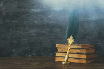 Old feather quill ink pen with inkwell and old books over wooden desk infront of black wall...