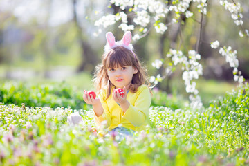 Cute little girl with curly hair wearing bunny ears and summer dress having fun during Easter egg hunt relaxing in the garden