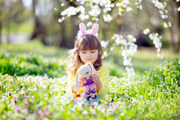 Cute little girl with curly hair wearing bunny ears and summer dress having fun during Easter egg hunt relaxing in the garden
