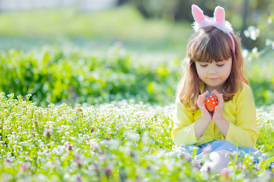 Cute little girl with curly hair wearing bunny ears and summer dress having fun during Easter egg hunt relaxing in the garden