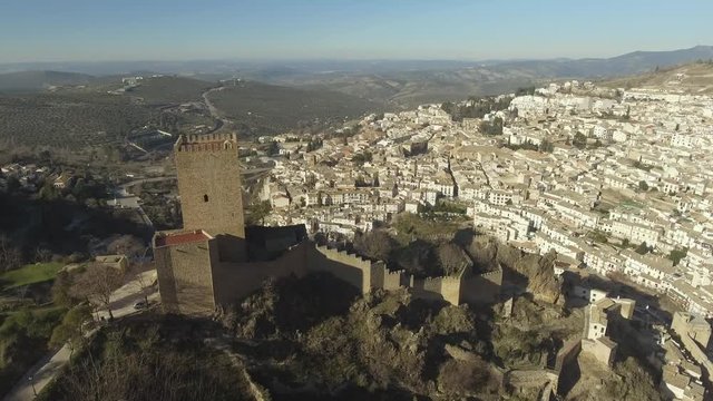 Aerial shot of Yerda Castle in gothic style on a steep, rocky hill, Cazorla city with white houses at the foot of the mountain and olive groves at the distance
