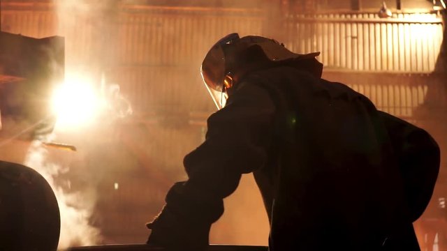Steel worker removing slag from the electric induction crucible melting furnace at the metallurgical plant, hard work conceprt. Stock footage. Man in protective mask and uniform working with a poker.