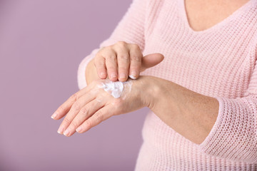 Mature woman applying hand cream on color background, closeup
