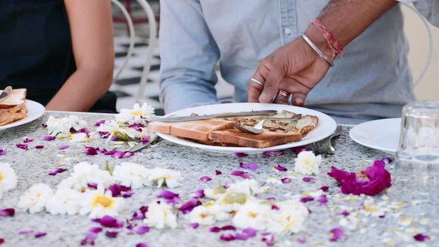 Waiter brings breakfast toasts and pancakes to a couple sitting at the table covered with flowers petals in hotel. Table close-up. Slow motion.