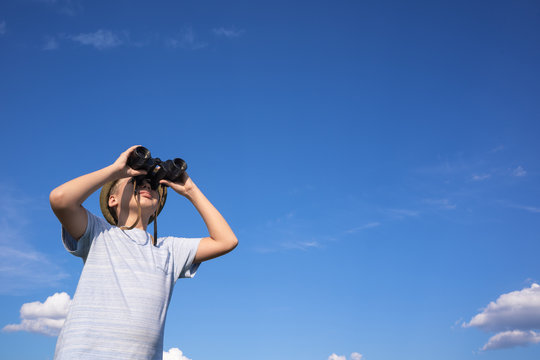 Side view of white kid looking through old vintage black binoculars at something far away in distance. Boy isolated on sunny clear blue sky background. 