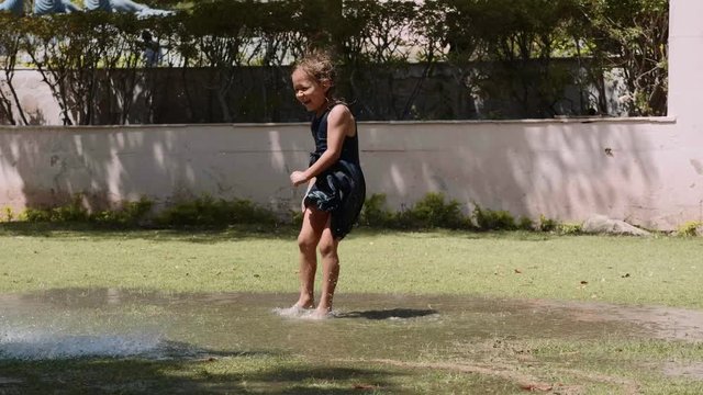 Happy little wet girl in dress is jumping in puddles on the lawn and making acrobatic wheel. Summer outdoor activities. Slow motion.