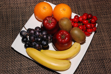 Fresh Oranges, apples, grapes, kiwis, cherries, bananas on the white plate on the brown table