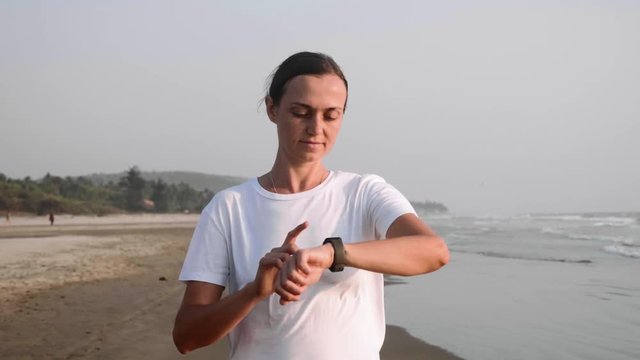 Young woman in white t-shirt is jogging on the ocean beach and starting fitness watch timer touching the screen, front view. Slow motion.