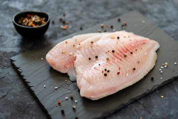Fresh tilapia fillet ready to be cooked, black wooden background. Side view, copy space