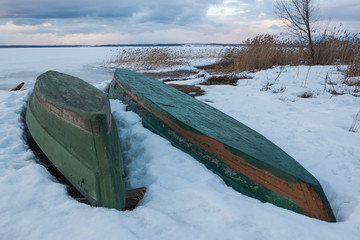 Old wooden boats lie in the snow on the shore of an ice-covered frozen lake in the winter. Close up.