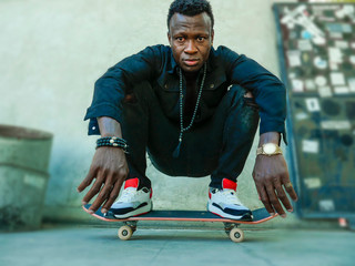 young attractive and serious black afro American man squatting on skate board at grunge street...
