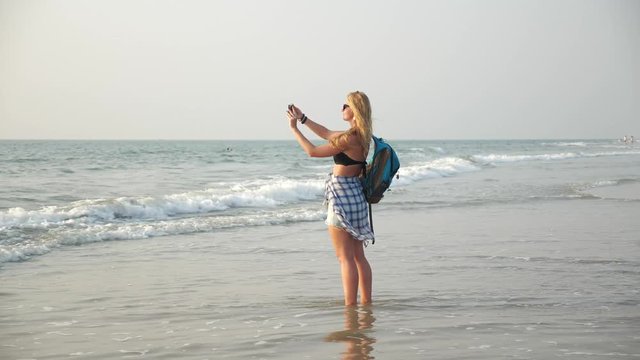 A young beautiful long-haired blonde girl with a backpack stands barefoot in the water of the sea on a sandy beach and takes pictures on a smartphone and selfie. A woman walks by the ocean on vacation