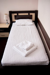Stack of white clean bath towels on bed sheet in modern hotel bedroom interior, copy space. Empty single bed with wooden headboard and bedside table