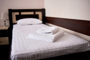 Stack of white clean bath towels on bed sheet in modern hotel bedroom interior, copy space. Empty single bed with wooden headboard and bedside table