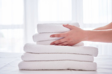 Woman putting stack of fresh white bath clean towels on bed sheet, copy space. Close up hands of hotel maid with towels. Room service