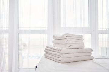 Stack of fresh white bath towels on bed sheet in modern hotel bedroom interior with window on...
