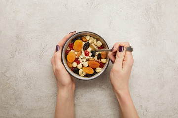 cropped view of woman holding spoon and bowl with muesli, dried apricots and berries, nuts on...