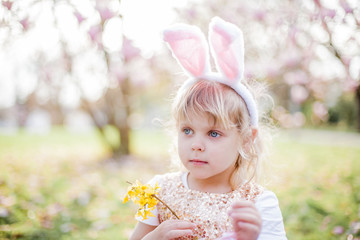 Little cute girl sitting on the grass near the magnolia. A girl dressed as an Easter bunny holds a flower and an egg.