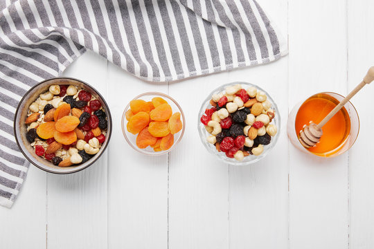 Top View Of Bowls With Cereal, Dried Apricots And Berries, Honey And Nuts On White Table With Striped Cloth