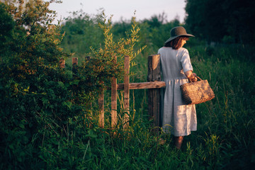 Stylish girl in linen dress holding rustic straw basket at wooden fence  in sunset light. Boho...