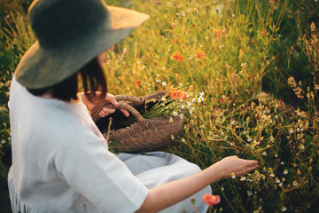 Stylish girl in linen dress gathering flowers in rustic straw basket, sitting in poppy meadow in sunset. Boho woman in hat relaxing in warm evening sunlight in summer field. Space for text