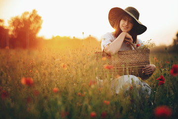 Stylish girl in linen dress smiling in poppy meadow in sunset light with flowers in rustic straw...