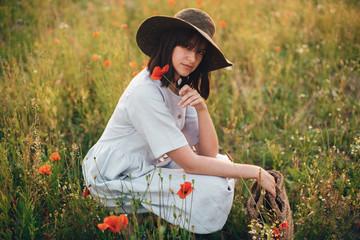 Stylish girl in linen dress holding poppy flower in meadow in sunset light with flowers in rustic...