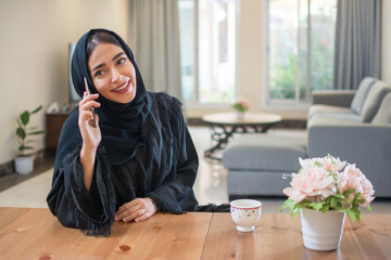 Smiling young muslim woman talking on mobile phone while enjoying morning coffee at home