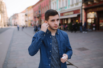 Man put in place his wireless headphones. Handsome man in blue shirt stand in centre if the city