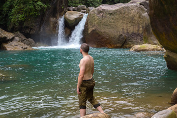 a man of beautiful physique and muscles with a naked body, stands against the background of a clean lake and a waterfall. travel and insurance.