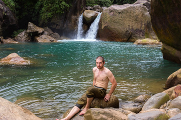 a man of beautiful body and muscles with a bare body, sits on the background of a clean lake and a waterfall because of a leg injury