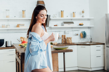 Smiling sexy girl in white lingerie holding cup of coffee in kitchen