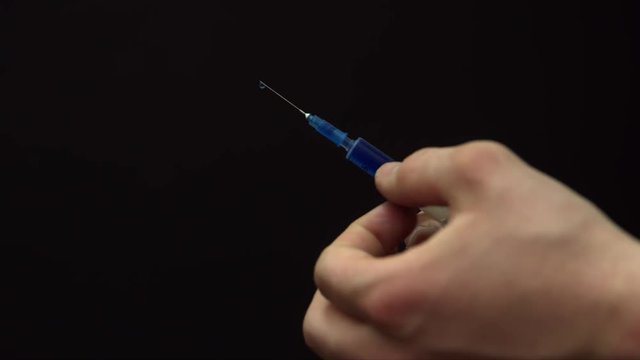 Hands of a young man tapping the syringe with a blue liquid inside, then he sprinkles some of the liquid into the air. Isolated on black.