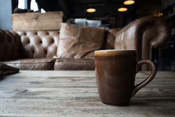 Tynemouth / Great Britain - April 6, 2019: Earthen ware mug cup on a rustic table with empty sofa behind.  Coffee or Tea Shop scene